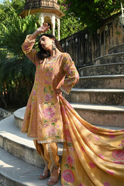 Model wearing a mustard yellow embroidered kurta set featuring intricate floral motifs in pink and green, delicate mirror and thread work, full sleeves, and a straight silhouette, paired with matching pants and a flowing floral dupatta, styled in an outdoor heritage garden setting.