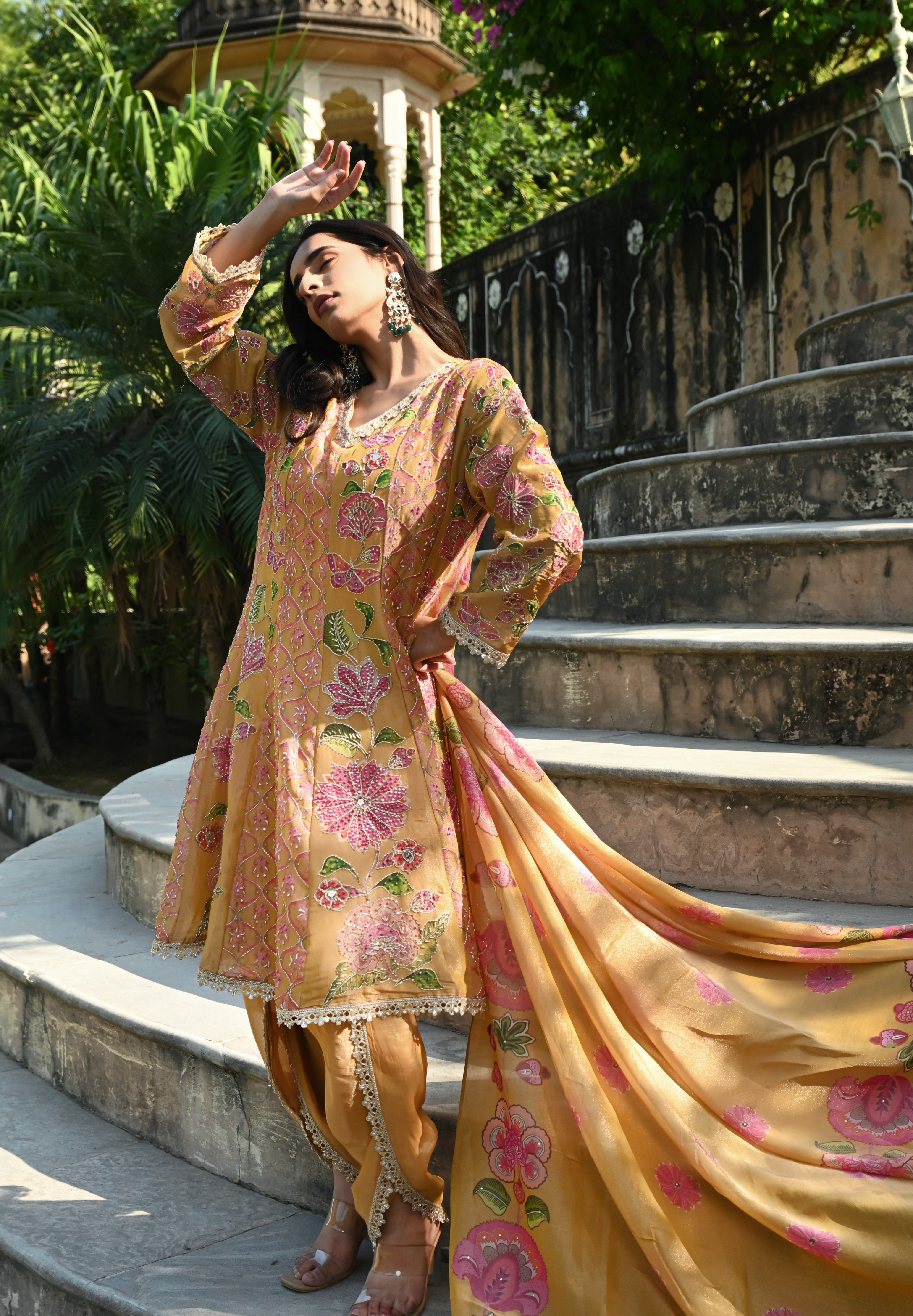 Model wearing a mustard yellow embroidered kurta set featuring intricate floral motifs in pink and green, delicate mirror and thread work, full sleeves, and a straight silhouette, paired with matching pants and a flowing floral dupatta, styled in an outdoor heritage garden setting.