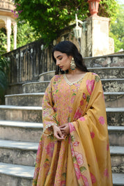 Model wearing a mustard yellow embroidered kurta set featuring intricate floral motifs in pink and green, delicate mirror and thread work, full sleeves, and a straight silhouette, paired with matching pants and a flowing floral dupatta, styled in an outdoor heritage garden setting.