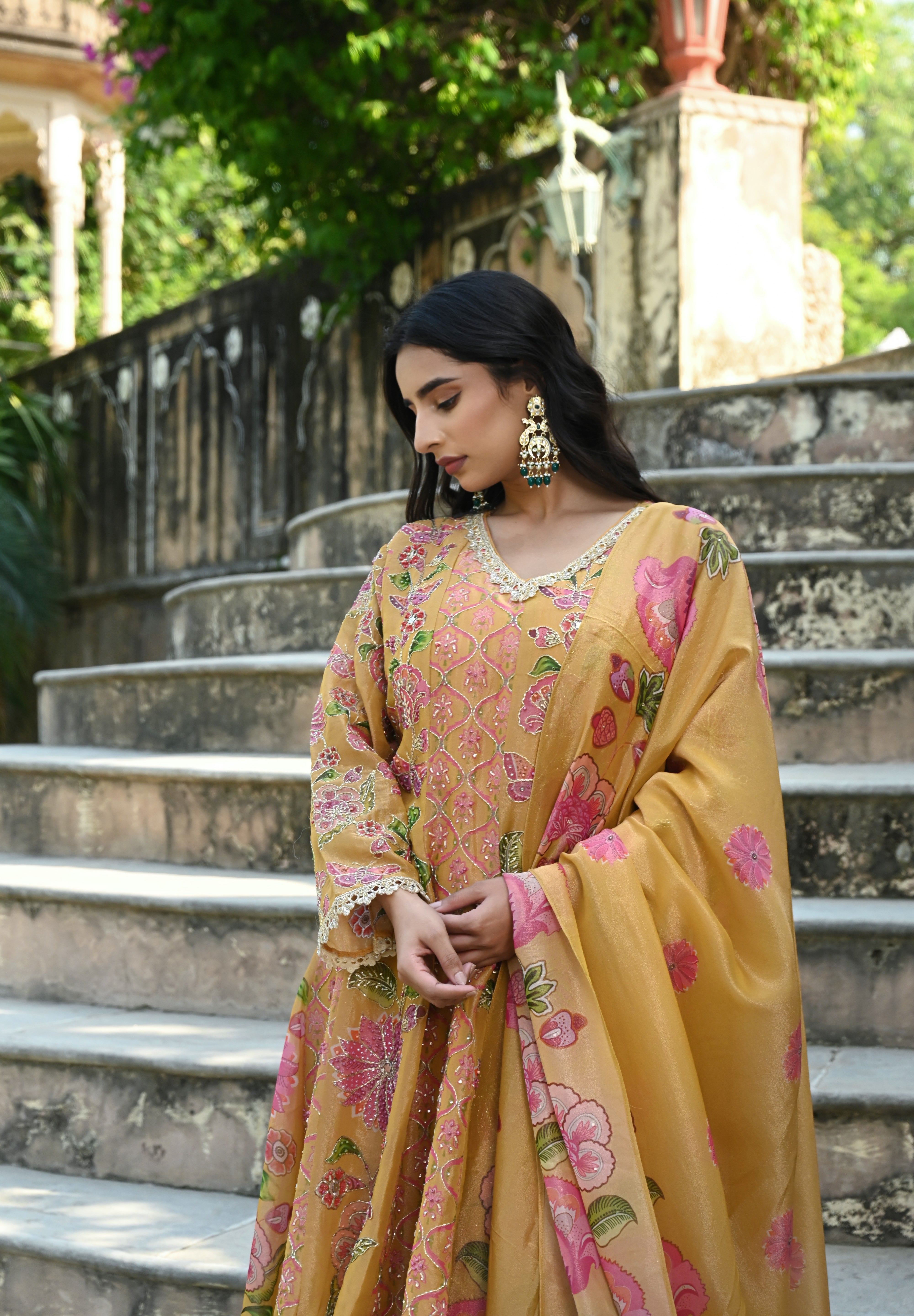 Model wearing a mustard yellow embroidered kurta set featuring intricate floral motifs in pink and green, delicate mirror and thread work, full sleeves, and a straight silhouette, paired with matching pants and a flowing floral dupatta, styled in an outdoor heritage garden setting.