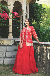 Model wearing a vibrant red embroidered lehenga set featuring a flowy skirt paired with a matching inner top and a short jacket-style blouse adorned with intricate mirror work and traditional motifs, styled with statement earrings in a lush heritage garden setting.
