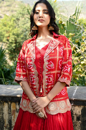 Model wearing a vibrant red embroidered lehenga set featuring a flowy skirt paired with a matching inner top and a short jacket-style blouse adorned with intricate mirror work and traditional motifs, styled with statement earrings in a lush heritage garden setting.