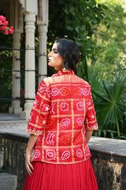Model wearing a vibrant red embroidered lehenga set featuring a flowy skirt paired with a matching inner top and a short jacket-style blouse adorned with intricate mirror work and traditional motifs, styled with statement earrings in a lush heritage garden setting.