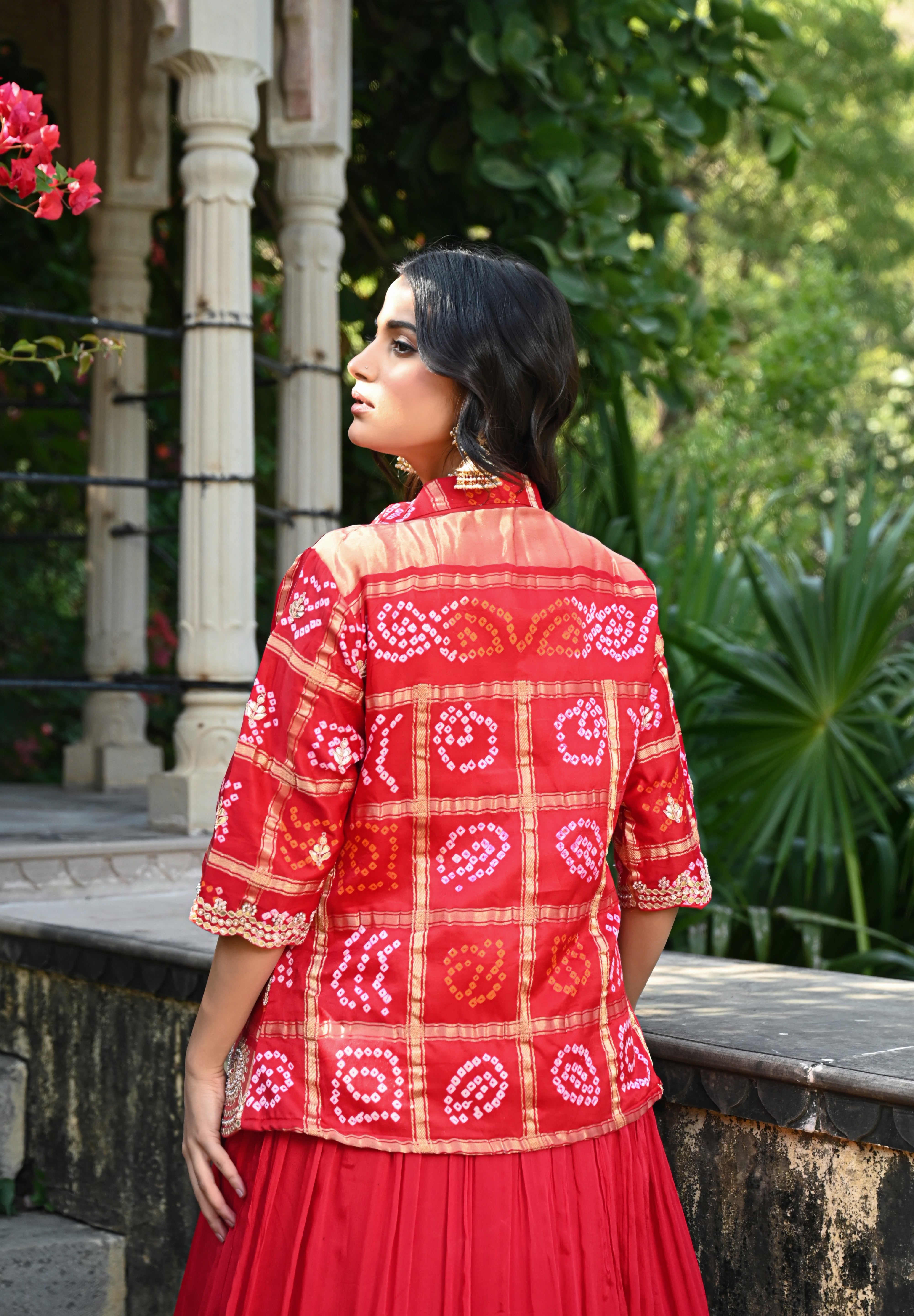 Model wearing a vibrant red embroidered lehenga set featuring a flowy skirt paired with a matching inner top and a short jacket-style blouse adorned with intricate mirror work and traditional motifs, styled with statement earrings in a lush heritage garden setting.