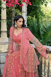Model wearing a pink printed co-ord set featuring a fitted blouse and wide-leg pants, layered with a long embroidered jacket adorned with intricate mirror work, tassel details, and traditional motifs, styled with statement earrings in a heritage garden setting.