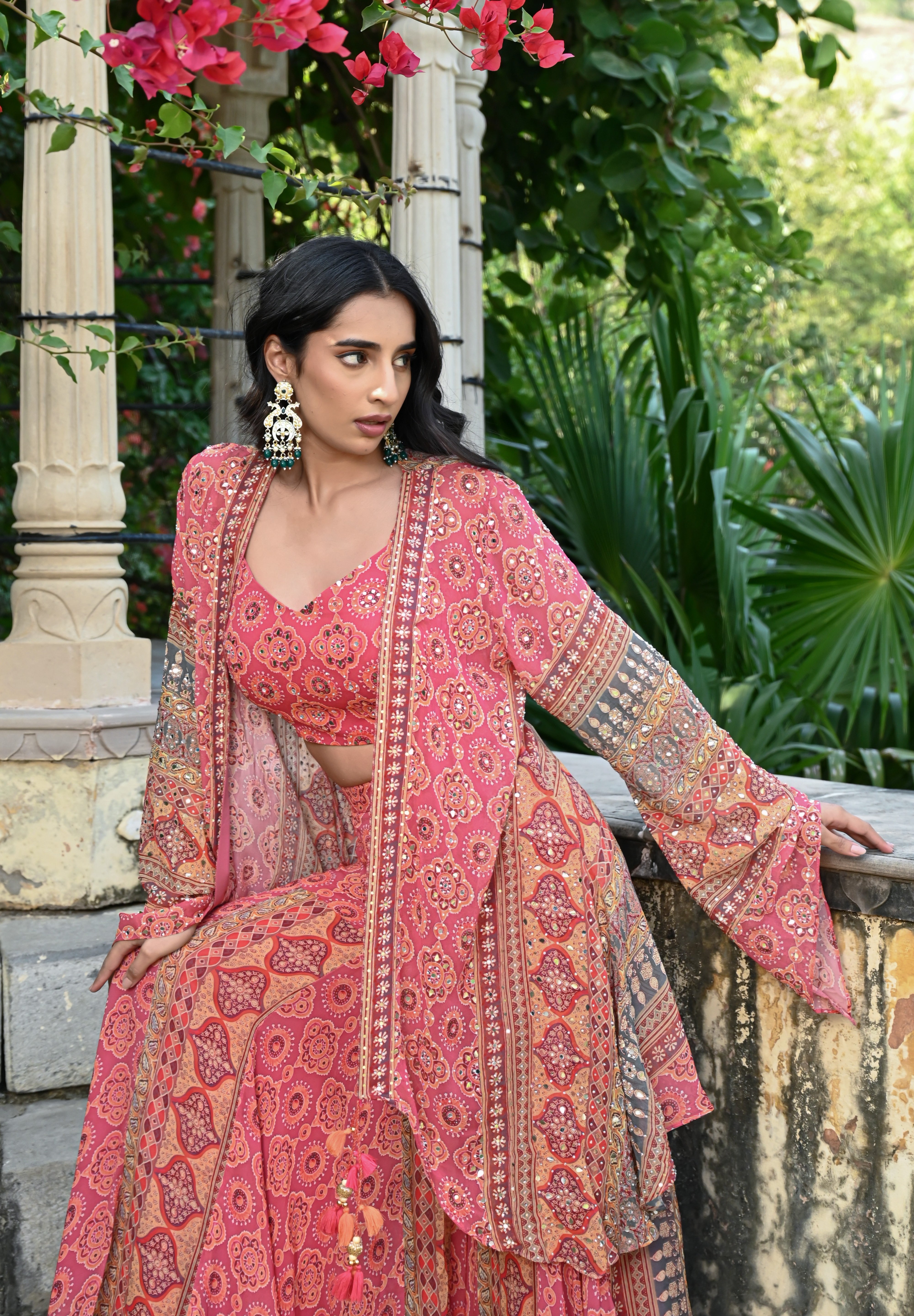 Model wearing a pink printed co-ord set featuring a fitted blouse and wide-leg pants, layered with a long embroidered jacket adorned with intricate mirror work, tassel details, and traditional motifs, styled with statement earrings in a heritage garden setting.