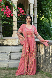Model wearing a pink printed co-ord set featuring a fitted blouse and wide-leg pants, layered with a long embroidered jacket adorned with intricate mirror work, tassel details, and traditional motifs, styled with statement earrings in a heritage garden setting.