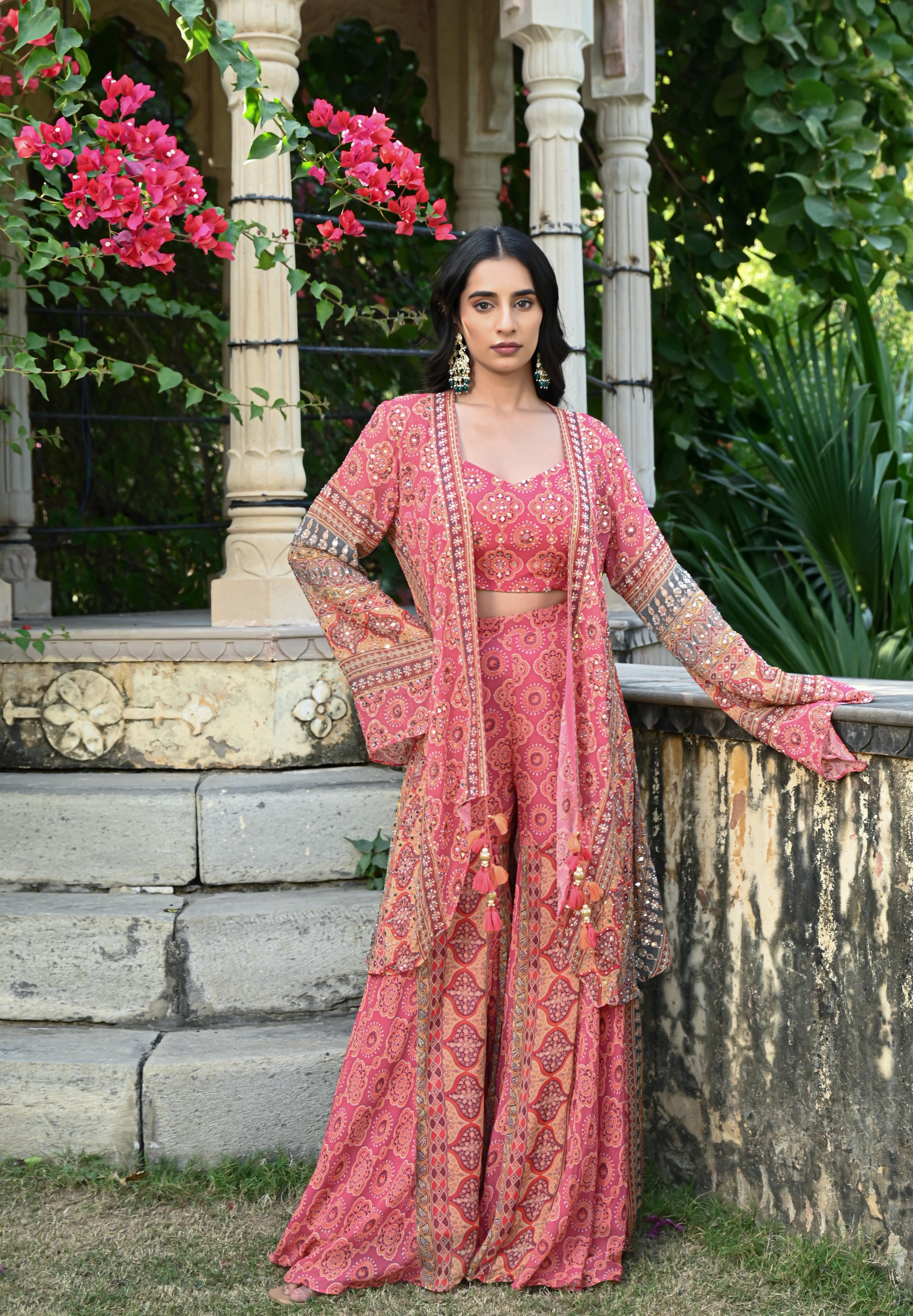 Model wearing a pink printed co-ord set featuring a fitted blouse and wide-leg pants, layered with a long embroidered jacket adorned with intricate mirror work, tassel details, and traditional motifs, styled with statement earrings in a heritage garden setting.