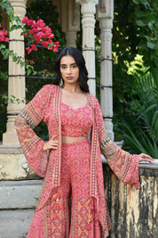 Model wearing a pink printed co-ord set featuring a fitted blouse and wide-leg pants, layered with a long embroidered jacket adorned with intricate mirror work, tassel details, and traditional motifs, styled with statement earrings in a heritage garden setting.