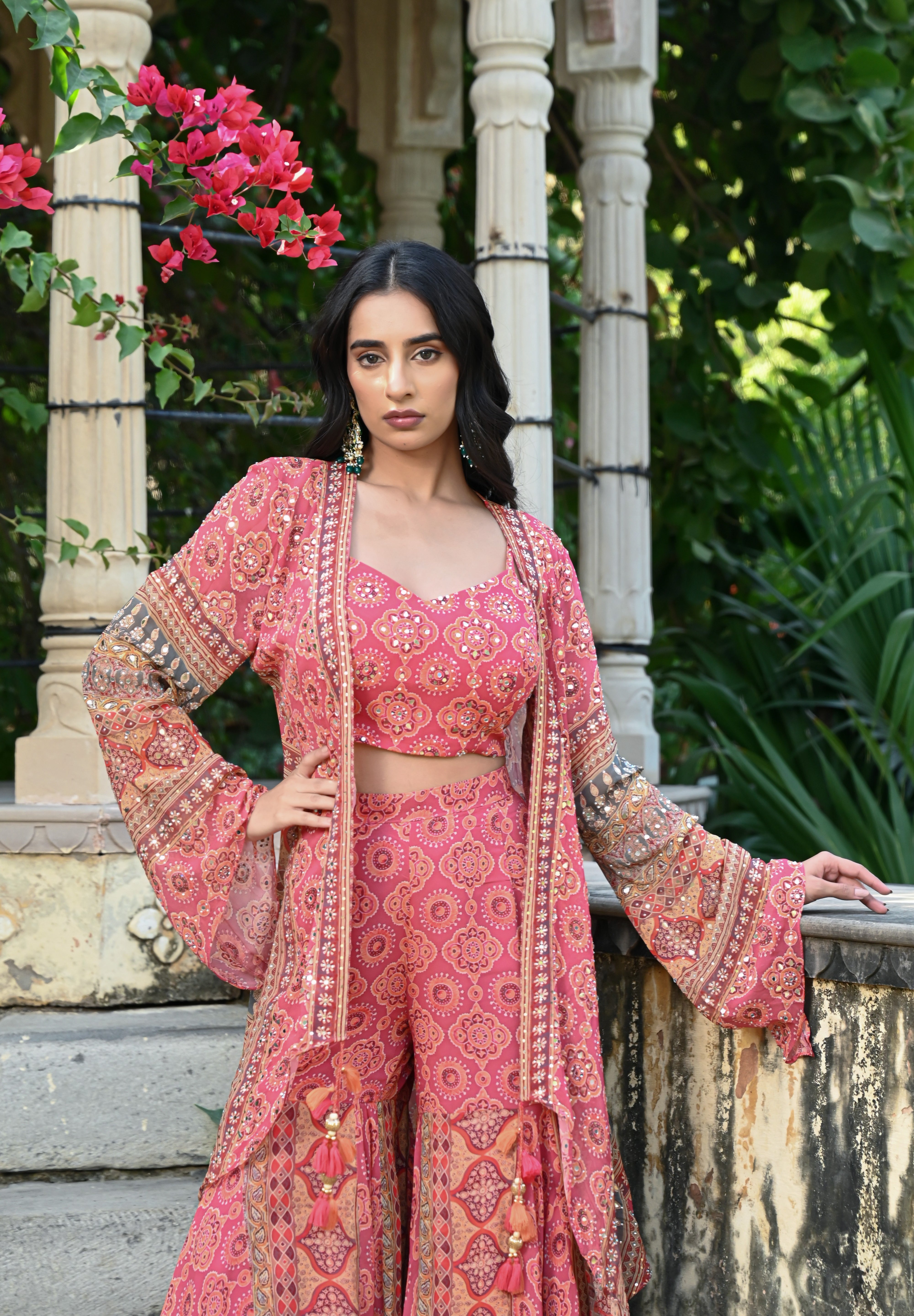 Model wearing a pink printed co-ord set featuring a fitted blouse and wide-leg pants, layered with a long embroidered jacket adorned with intricate mirror work, tassel details, and traditional motifs, styled with statement earrings in a heritage garden setting.
