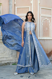 A woman stands posing in a dusty blue traditional Indian ensemble against a rustic, peach-colored wall with architectural arches. The outfit features a long, sleeveless front-slit jacket adorned with intricate silver floral and paisley embroidery, paired with matching wide-leg metallic trousers and a draped dupatta. She completes the look with large silver statement earrings and silver pointed-toe flats with bow detailing.