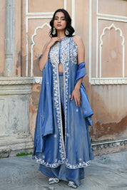 A woman stands posing in a dusty blue traditional Indian ensemble against a rustic, peach-colored wall with architectural arches. The outfit features a long, sleeveless front-slit jacket adorned with intricate silver floral and paisley embroidery, paired with matching wide-leg metallic trousers and a draped dupatta. She completes the look with large silver statement earrings and silver pointed-toe flats with bow detailing.