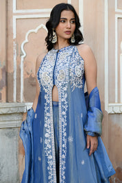 A woman stands posing in a dusty blue traditional Indian ensemble against a rustic, peach-colored wall with architectural arches. The outfit features a long, sleeveless front-slit jacket adorned with intricate silver floral and paisley embroidery, paired with matching wide-leg metallic trousers and a draped dupatta. She completes the look with large silver statement earrings and silver pointed-toe flats with bow detailing.