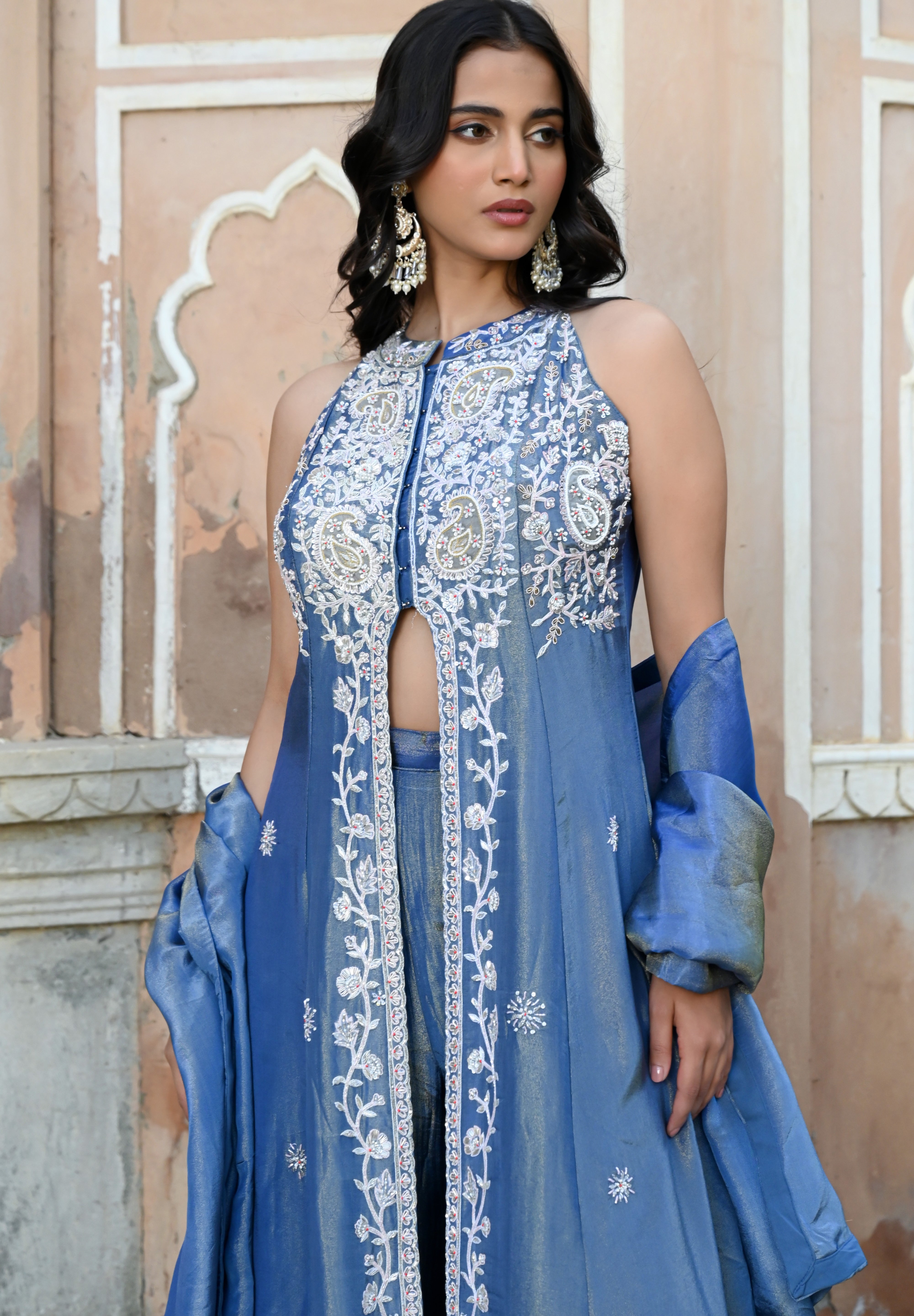 A woman stands posing in a dusty blue traditional Indian ensemble against a rustic, peach-colored wall with architectural arches. The outfit features a long, sleeveless front-slit jacket adorned with intricate silver floral and paisley embroidery, paired with matching wide-leg metallic trousers and a draped dupatta. She completes the look with large silver statement earrings and silver pointed-toe flats with bow detailing.