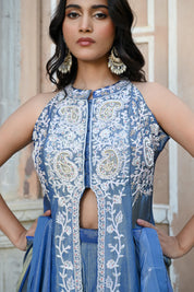 A woman stands posing in a dusty blue traditional Indian ensemble against a rustic, peach-colored wall with architectural arches. The outfit features a long, sleeveless front-slit jacket adorned with intricate silver floral and paisley embroidery, paired with matching wide-leg metallic trousers and a draped dupatta. She completes the look with large silver statement earrings and silver pointed-toe flats with bow detailing.
