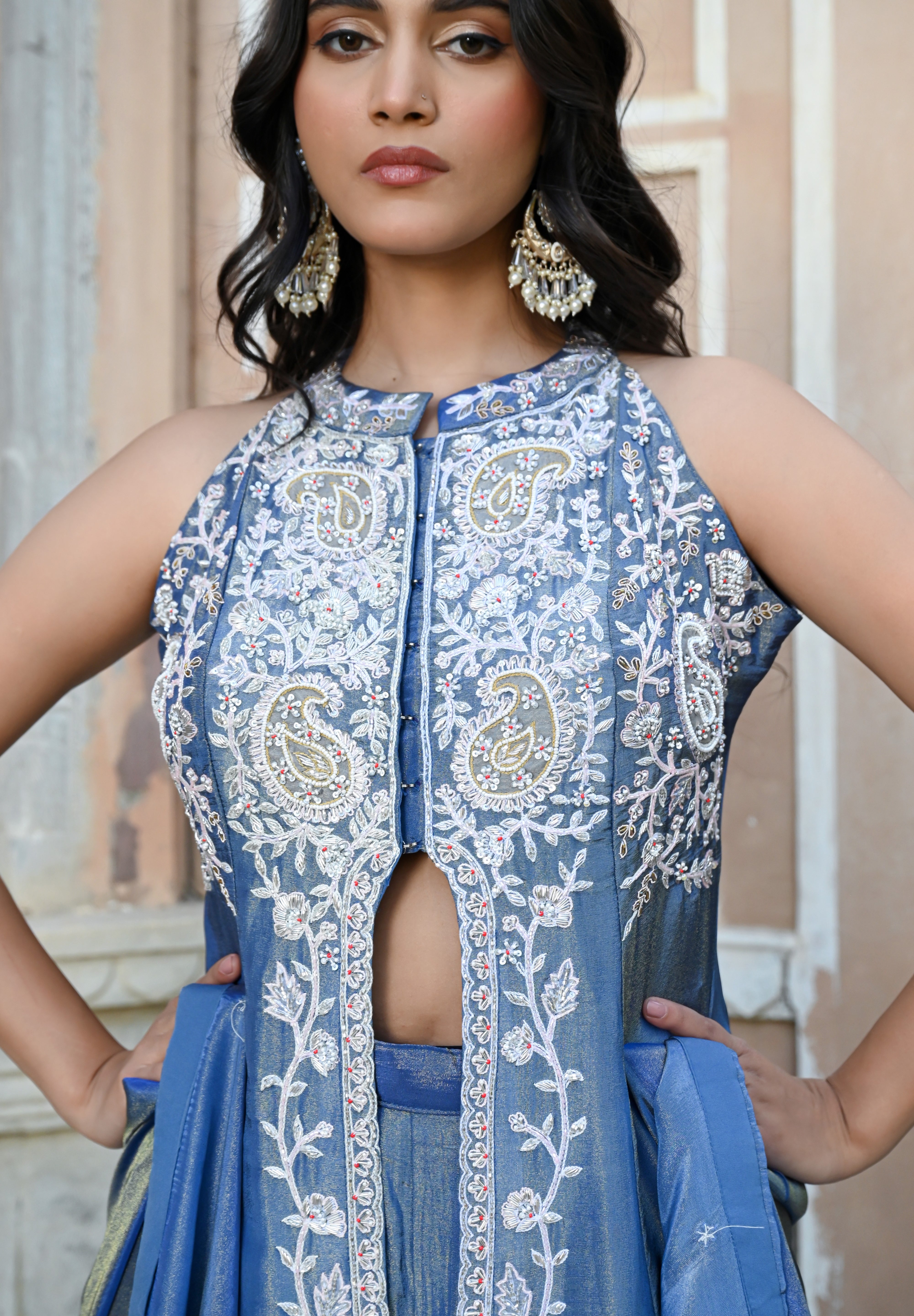 A woman stands posing in a dusty blue traditional Indian ensemble against a rustic, peach-colored wall with architectural arches. The outfit features a long, sleeveless front-slit jacket adorned with intricate silver floral and paisley embroidery, paired with matching wide-leg metallic trousers and a draped dupatta. She completes the look with large silver statement earrings and silver pointed-toe flats with bow detailing.