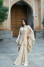 A woman with long, dark wavy hair poses in an elegant cream-colored Indo-western ensemble. She is wearing a collared tunic featuring intricate gold embroidery on the chest, paired with a matching sheer cape that has scalloped, lace-trimmed edges and a flowing satin skirt. She completes the look with long white statement earrings, standing in a stone-paved courtyard with a large rustic wooden door in the background.