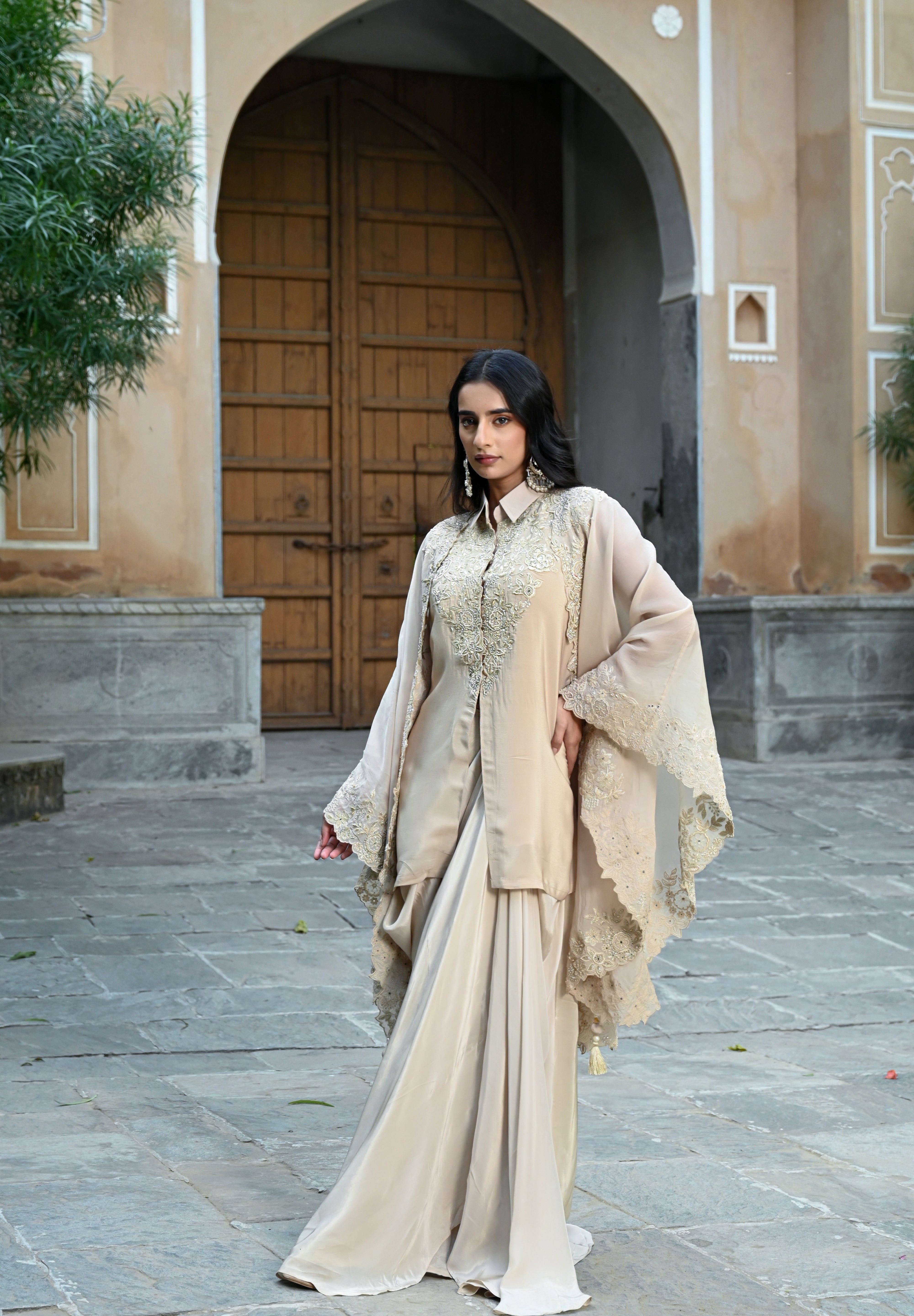 A woman with long, dark wavy hair poses in an elegant cream-colored Indo-western ensemble. She is wearing a collared tunic featuring intricate gold embroidery on the chest, paired with a matching sheer cape that has scalloped, lace-trimmed edges and a flowing satin skirt. She completes the look with long white statement earrings, standing in a stone-paved courtyard with a large rustic wooden door in the background.