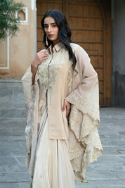 A woman with long, dark wavy hair poses in an elegant cream-colored Indo-western ensemble. She is wearing a collared tunic featuring intricate gold embroidery on the chest, paired with a matching sheer cape that has scalloped, lace-trimmed edges and a flowing satin skirt. She completes the look with long white statement earrings, standing in a stone-paved courtyard with a large rustic wooden door in the background.