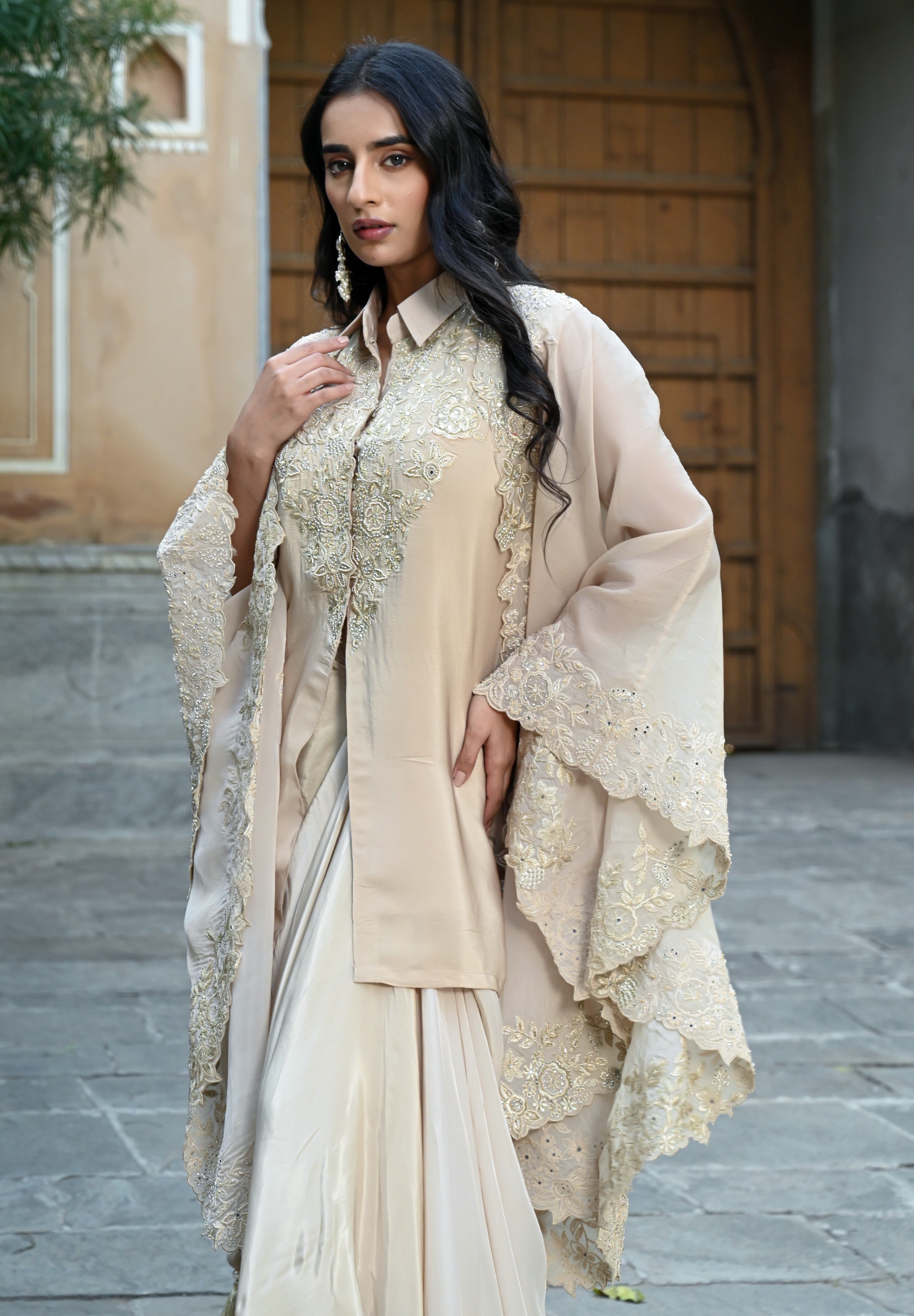 A woman with long, dark wavy hair poses in an elegant cream-colored Indo-western ensemble. She is wearing a collared tunic featuring intricate gold embroidery on the chest, paired with a matching sheer cape that has scalloped, lace-trimmed edges and a flowing satin skirt. She completes the look with long white statement earrings, standing in a stone-paved courtyard with a large rustic wooden door in the background.
