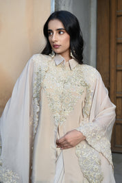 A woman with long, dark wavy hair poses in an elegant cream-colored Indo-western ensemble. She is wearing a collared tunic featuring intricate gold embroidery on the chest, paired with a matching sheer cape that has scalloped, lace-trimmed edges and a flowing satin skirt. She completes the look with long white statement earrings, standing in a stone-paved courtyard with a large rustic wooden door in the background.