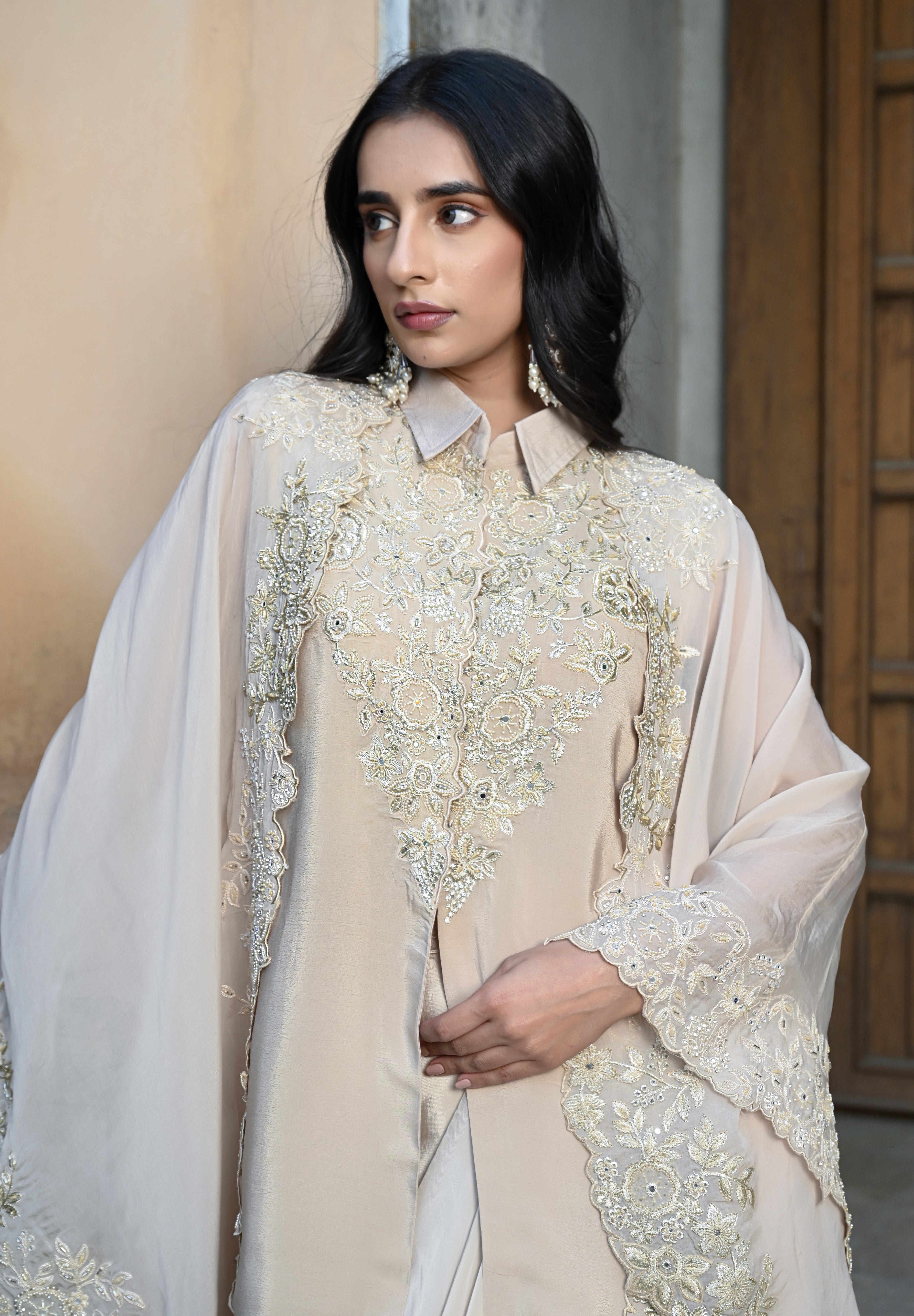 A woman with long, dark wavy hair poses in an elegant cream-colored Indo-western ensemble. She is wearing a collared tunic featuring intricate gold embroidery on the chest, paired with a matching sheer cape that has scalloped, lace-trimmed edges and a flowing satin skirt. She completes the look with long white statement earrings, standing in a stone-paved courtyard with a large rustic wooden door in the background.