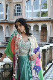 A woman with long, dark wavy hair poses in an elegant cream-colored Indo-western ensemble. She wears a collared tunic featuring intricate gold embroidery on the chest, paired with a matching sheer cape that has scalloped, embroidered edges and a flowing satin skirt. She stands in a stone-paved courtyard with a large rustic wooden door and traditional arched architecture in the background.