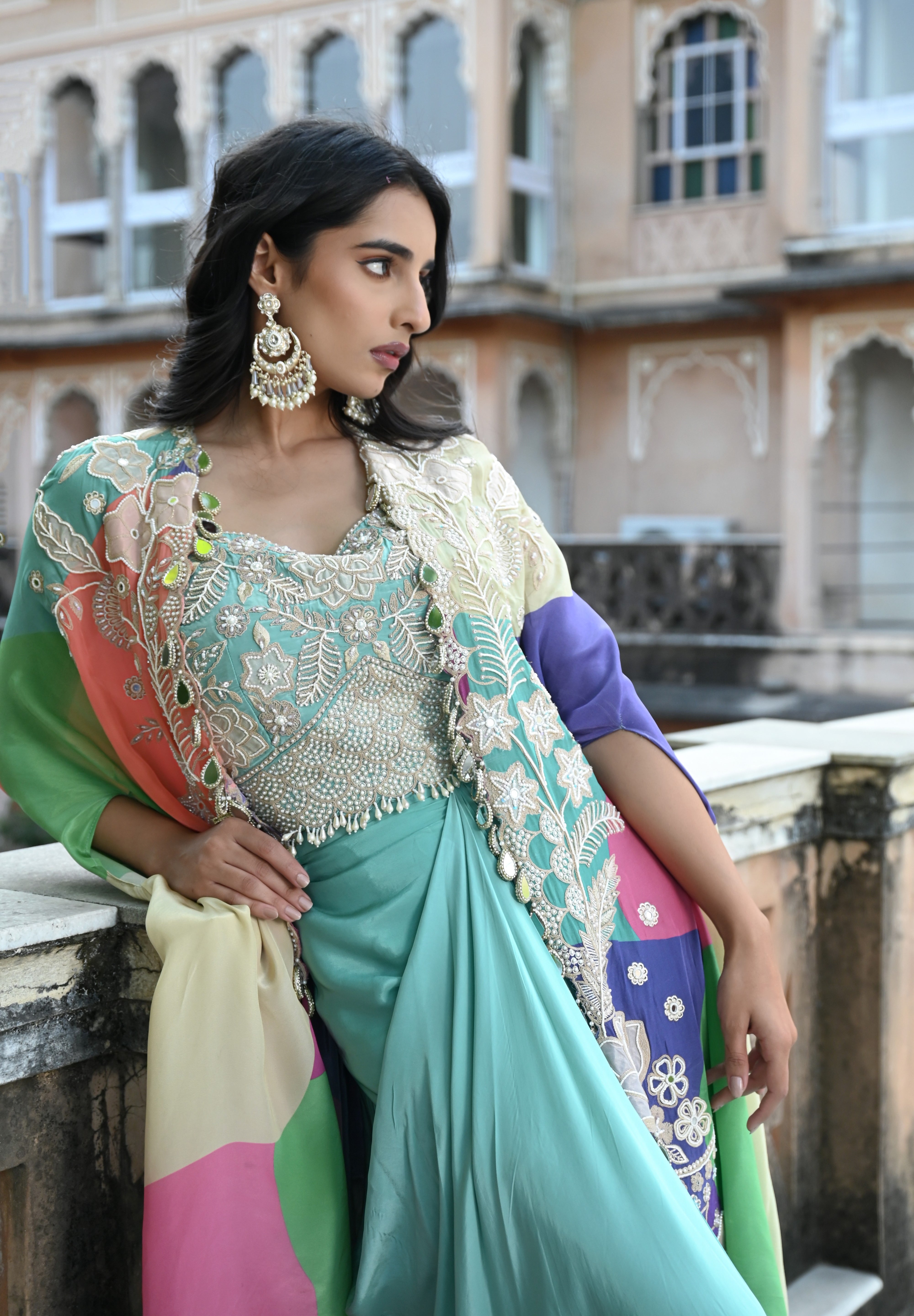 A woman with long, dark wavy hair poses in an elegant cream-colored Indo-western ensemble. She wears a collared tunic featuring intricate gold embroidery on the chest, paired with a matching sheer cape that has scalloped, embroidered edges and a flowing satin skirt. She stands in a stone-paved courtyard with a large rustic wooden door and traditional arched architecture in the background.