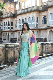 A woman with long, dark wavy hair poses in an elegant cream-colored Indo-western ensemble. She wears a collared tunic featuring intricate gold embroidery on the chest, paired with a matching sheer cape that has scalloped, embroidered edges and a flowing satin skirt. She stands in a stone-paved courtyard with a large rustic wooden door and traditional arched architecture in the background.