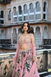 A woman with long, dark wavy hair poses in an elegant cream-colored Indo-western ensemble. She wears a collared tunic featuring intricate gold embroidery on the chest, paired with a matching sheer cape that has scalloped, embroidered edges and a flowing satin skirt. She stands in a stone-paved courtyard with a large rustic wooden door and traditional arched architecture in the background.