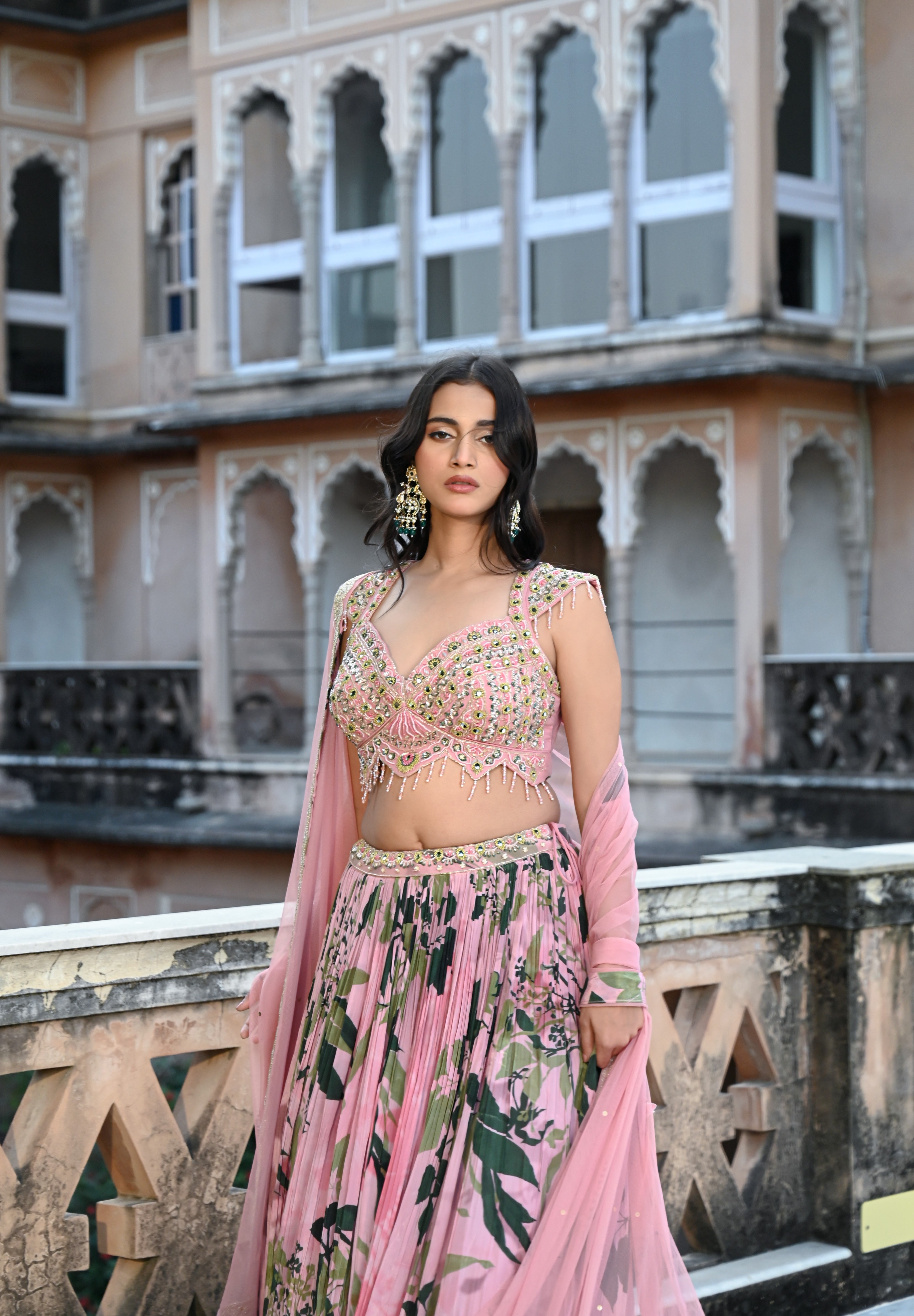 A woman with long, dark wavy hair poses in an elegant cream-colored Indo-western ensemble. She wears a collared tunic featuring intricate gold embroidery on the chest, paired with a matching sheer cape that has scalloped, embroidered edges and a flowing satin skirt. She stands in a stone-paved courtyard with a large rustic wooden door and traditional arched architecture in the background.