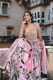 A woman with long, dark wavy hair poses in an elegant cream-colored Indo-western ensemble. She wears a collared tunic featuring intricate gold embroidery on the chest, paired with a matching sheer cape that has scalloped, embroidered edges and a flowing satin skirt. She stands in a stone-paved courtyard with a large rustic wooden door and traditional arched architecture in the background.