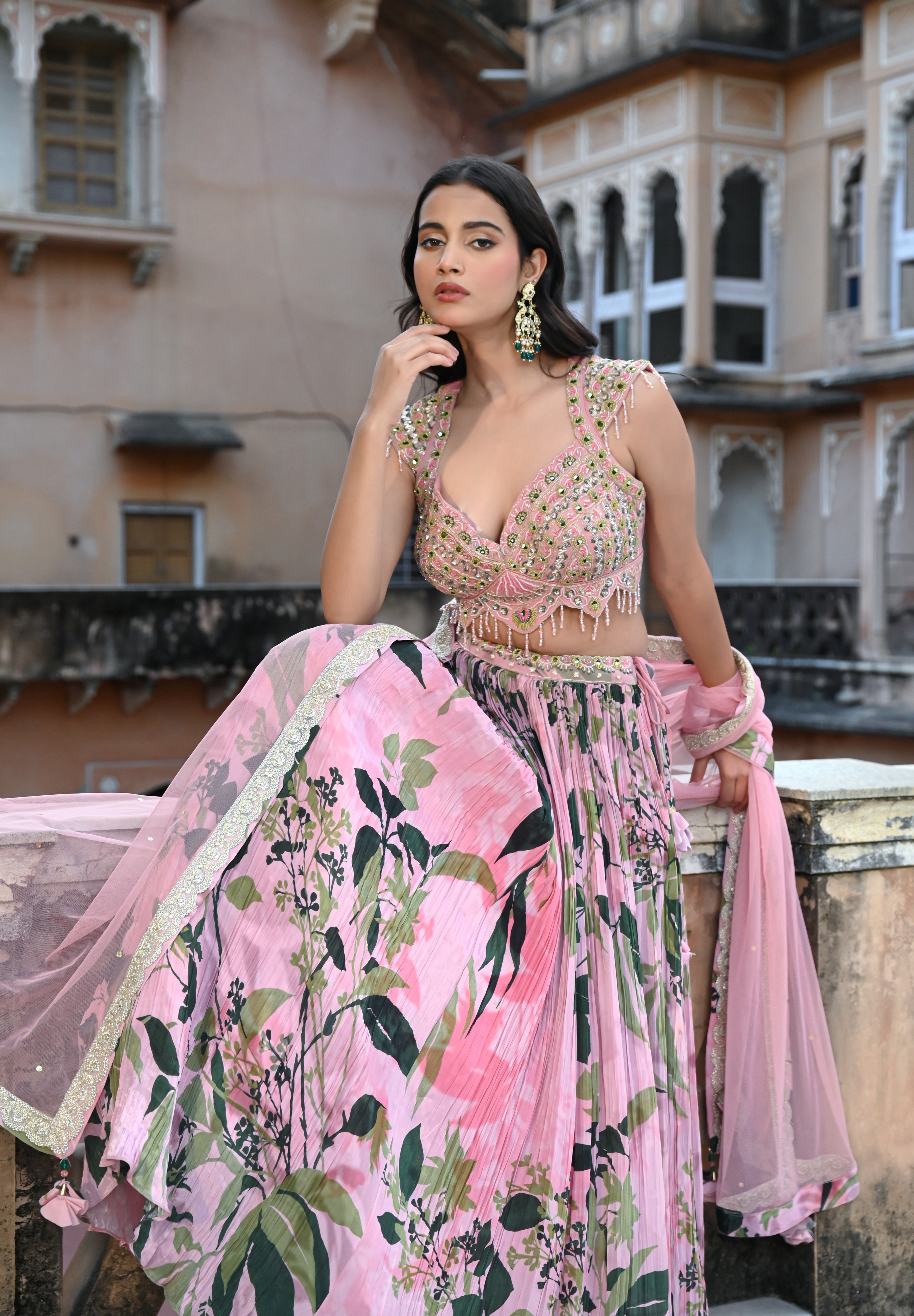 A woman with long, dark wavy hair poses in an elegant cream-colored Indo-western ensemble. She wears a collared tunic featuring intricate gold embroidery on the chest, paired with a matching sheer cape that has scalloped, embroidered edges and a flowing satin skirt. She stands in a stone-paved courtyard with a large rustic wooden door and traditional arched architecture in the background.