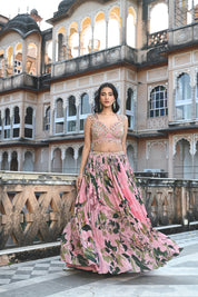 A woman with long, dark wavy hair poses in an elegant cream-colored Indo-western ensemble. She wears a collared tunic featuring intricate gold embroidery on the chest, paired with a matching sheer cape that has scalloped, embroidered edges and a flowing satin skirt. She stands in a stone-paved courtyard with a large rustic wooden door and traditional arched architecture in the background.