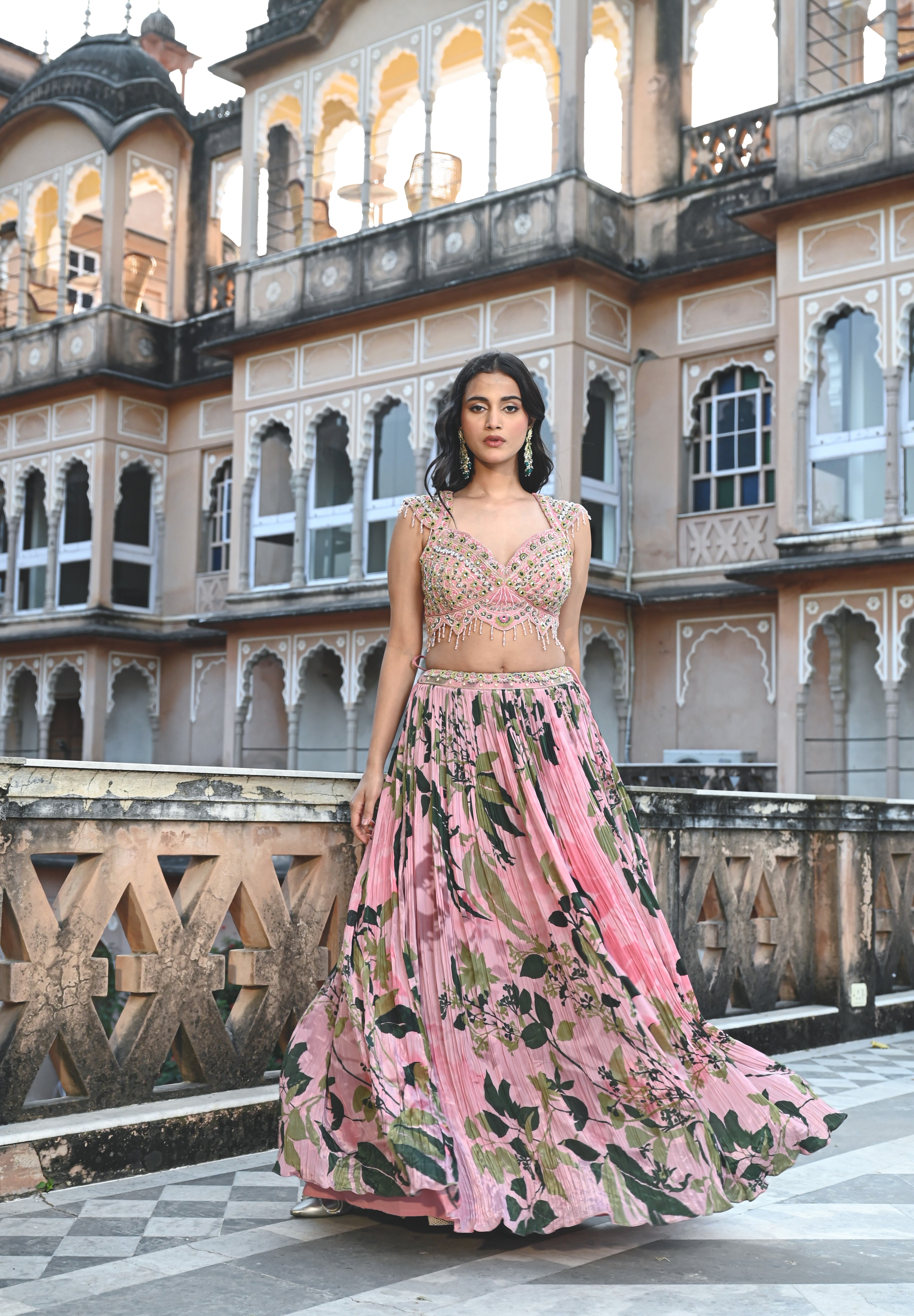 A woman with long, dark wavy hair poses in an elegant cream-colored Indo-western ensemble. She wears a collared tunic featuring intricate gold embroidery on the chest, paired with a matching sheer cape that has scalloped, embroidered edges and a flowing satin skirt. She stands in a stone-paved courtyard with a large rustic wooden door and traditional arched architecture in the background.