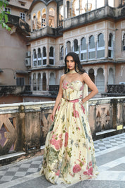A woman stands on a rooftop terrace wearing a cream-colored lehenga set with a romantic floral print in shades of pink and green. The outfit includes a sweetheart neckline blouse with spaghetti straps and a voluminous pleated skirt. She is accessorized with large gold statement earrings and stands against a backdrop of a historic Indian building featuring numerous decorative arched balconies and windows.