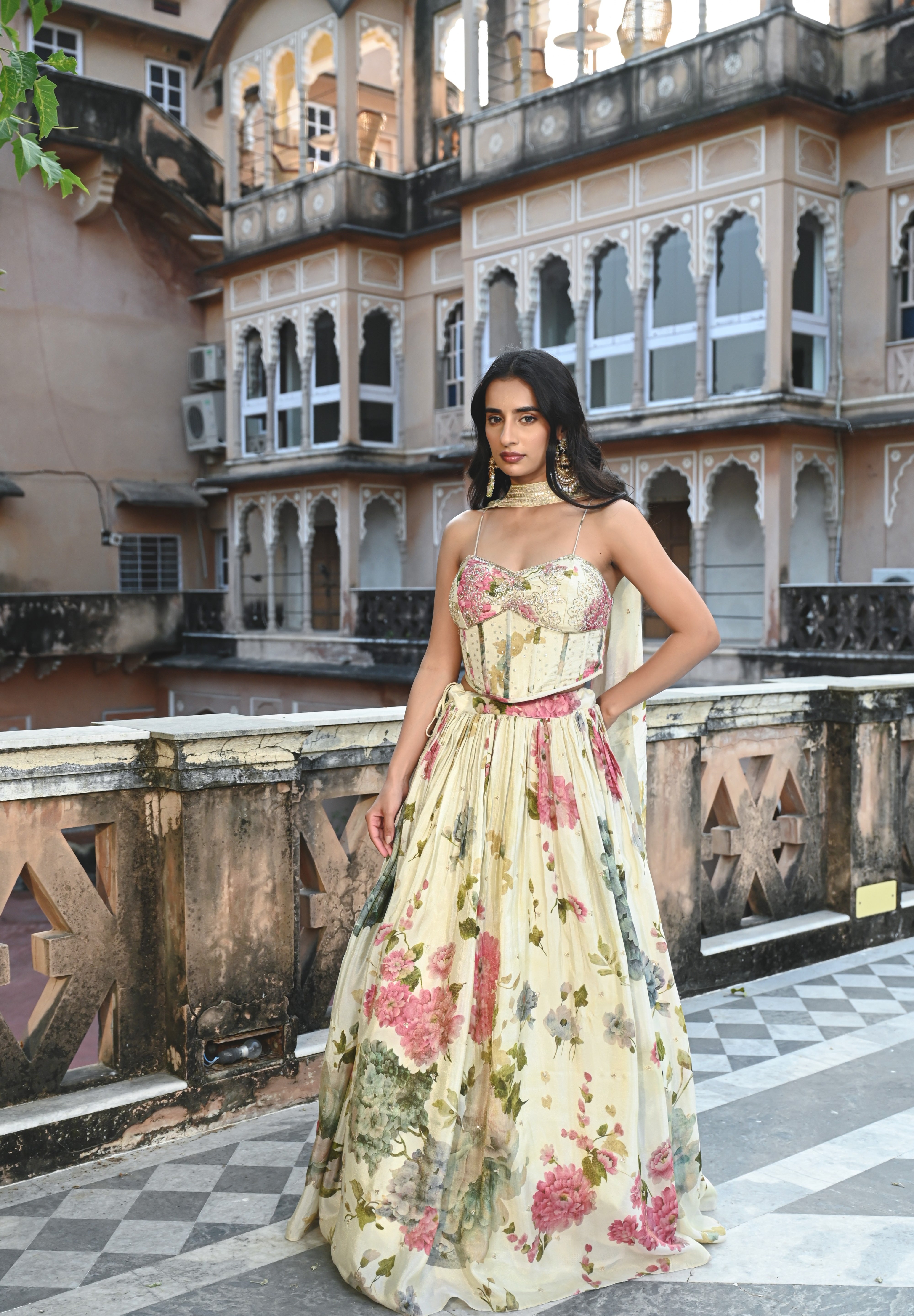 A woman stands on a rooftop terrace wearing a cream-colored lehenga set with a romantic floral print in shades of pink and green. The outfit includes a sweetheart neckline blouse with spaghetti straps and a voluminous pleated skirt. She is accessorized with large gold statement earrings and stands against a backdrop of a historic Indian building featuring numerous decorative arched balconies and windows.