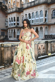A woman stands on a rooftop terrace wearing a cream-colored lehenga set with a romantic floral print in shades of pink and green. The outfit includes a sweetheart neckline blouse with spaghetti straps and a voluminous pleated skirt. She is accessorized with large gold statement earrings and stands against a backdrop of a historic Indian building featuring numerous decorative arched balconies and windows.