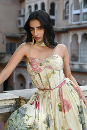 A woman stands on a rooftop terrace wearing a cream-colored lehenga set with a romantic floral print in shades of pink and green. The outfit includes a sweetheart neckline blouse with spaghetti straps and a voluminous pleated skirt. She is accessorized with large gold statement earrings and stands against a backdrop of a historic Indian building featuring numerous decorative arched balconies and windows.