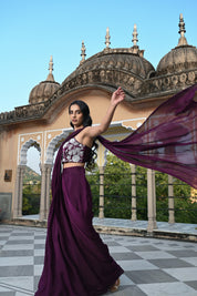 A woman poses on a rooftop in a deep plum-colored draped saree-style outfit. She wears a sleeveless crop top with striking white floral embroidery, paired with a flowing skirt and a long sheer fabric trailing behind her in the wind. The setting features a clear blue sky and a historic building with traditional dome-shaped chhatris and arched pillars.