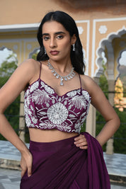 A woman poses on a rooftop in a deep plum-colored draped saree-style outfit. She wears a sleeveless crop top with striking white floral embroidery, paired with a flowing skirt and a long sheer fabric trailing behind her in the wind. The setting features a clear blue sky and a historic building with traditional dome-shaped chhatris and arched pillars.