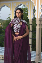 A woman poses on a rooftop in a deep plum-colored draped saree-style outfit. She wears a sleeveless crop top with striking white floral embroidery, paired with a flowing skirt and a long sheer fabric trailing behind her in the wind. The setting features a clear blue sky and a historic building with traditional dome-shaped chhatris and arched pillars.