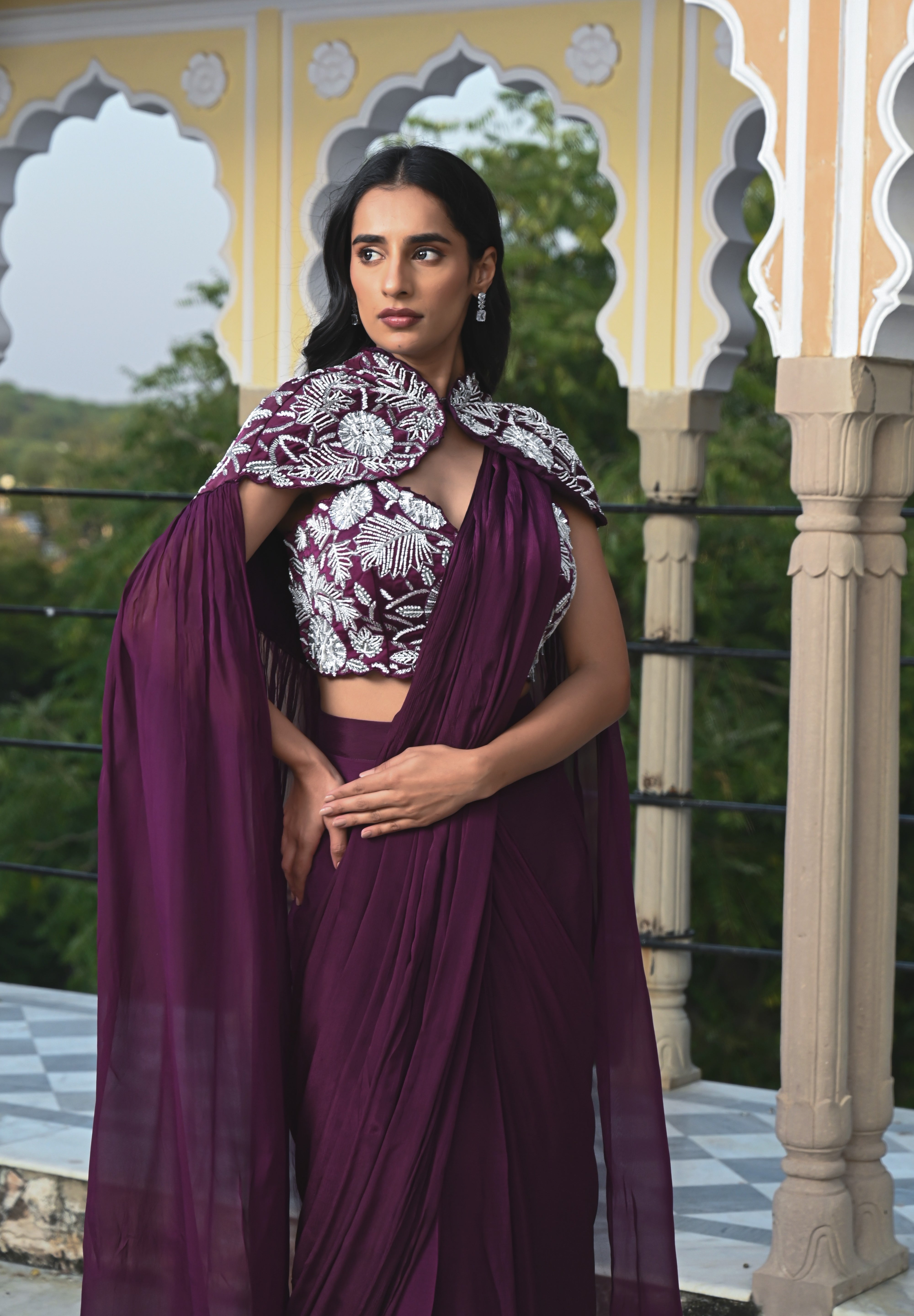 A woman poses on a rooftop in a deep plum-colored draped saree-style outfit. She wears a sleeveless crop top with striking white floral embroidery, paired with a flowing skirt and a long sheer fabric trailing behind her in the wind. The setting features a clear blue sky and a historic building with traditional dome-shaped chhatris and arched pillars.