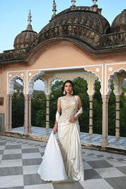 A woman poses on a rooftop in a shimmering ivory gown with a heavily embroidered bodice and a long, flowing train. The background features traditional Indian architectural elements like stone domes and scalloped arches.
