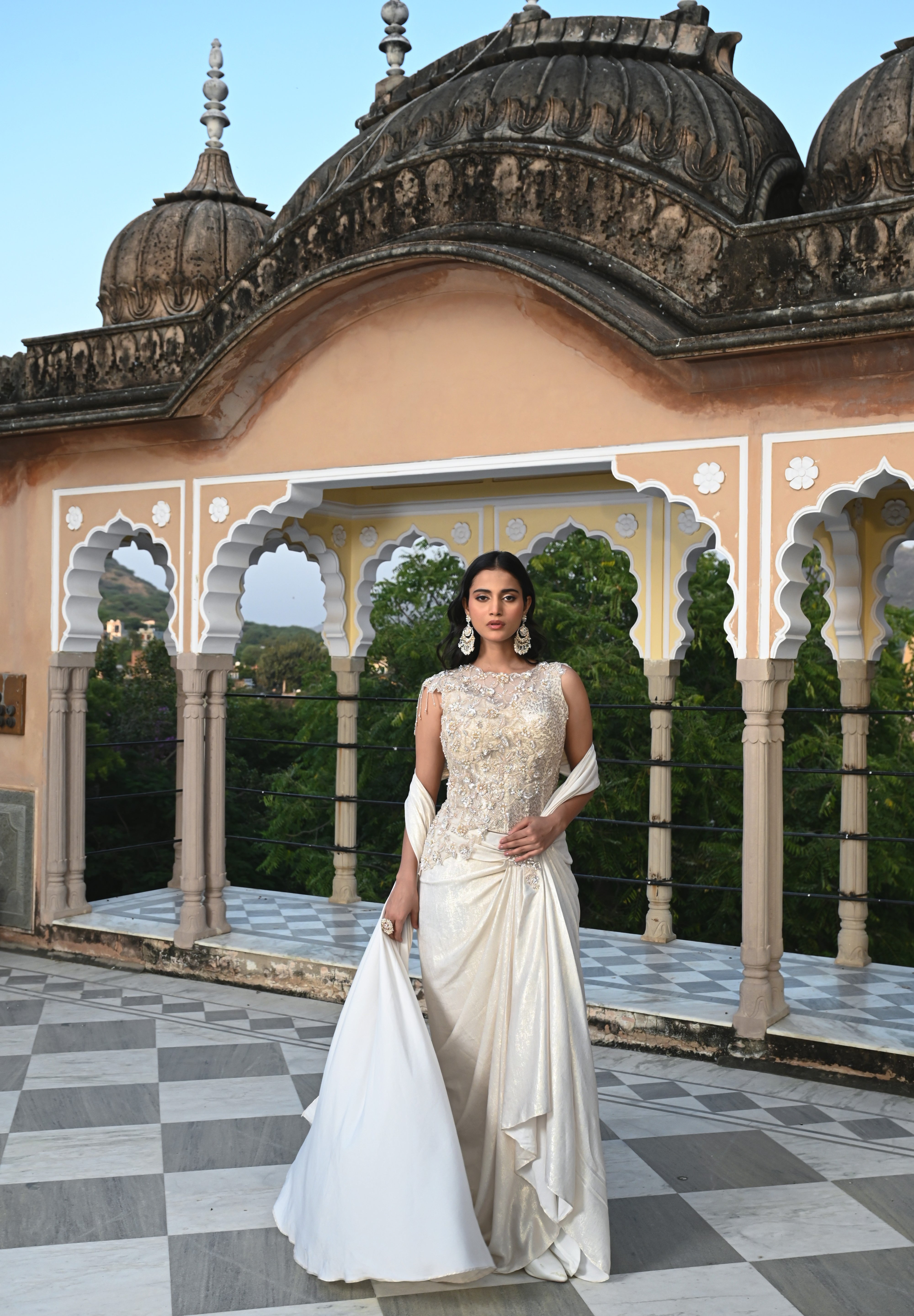 A woman poses on a rooftop in a shimmering ivory gown with a heavily embroidered bodice and a long, flowing train. The background features traditional Indian architectural elements like stone domes and scalloped arches.