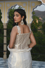 A woman poses on a rooftop in a shimmering ivory gown with a heavily embroidered bodice and a long, flowing train. The background features traditional Indian architectural elements like stone domes and scalloped arches.