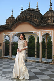 A woman poses on a rooftop in a shimmering ivory gown with a heavily embroidered bodice and a long, flowing train. The background features traditional Indian architectural elements like stone domes and scalloped arches.