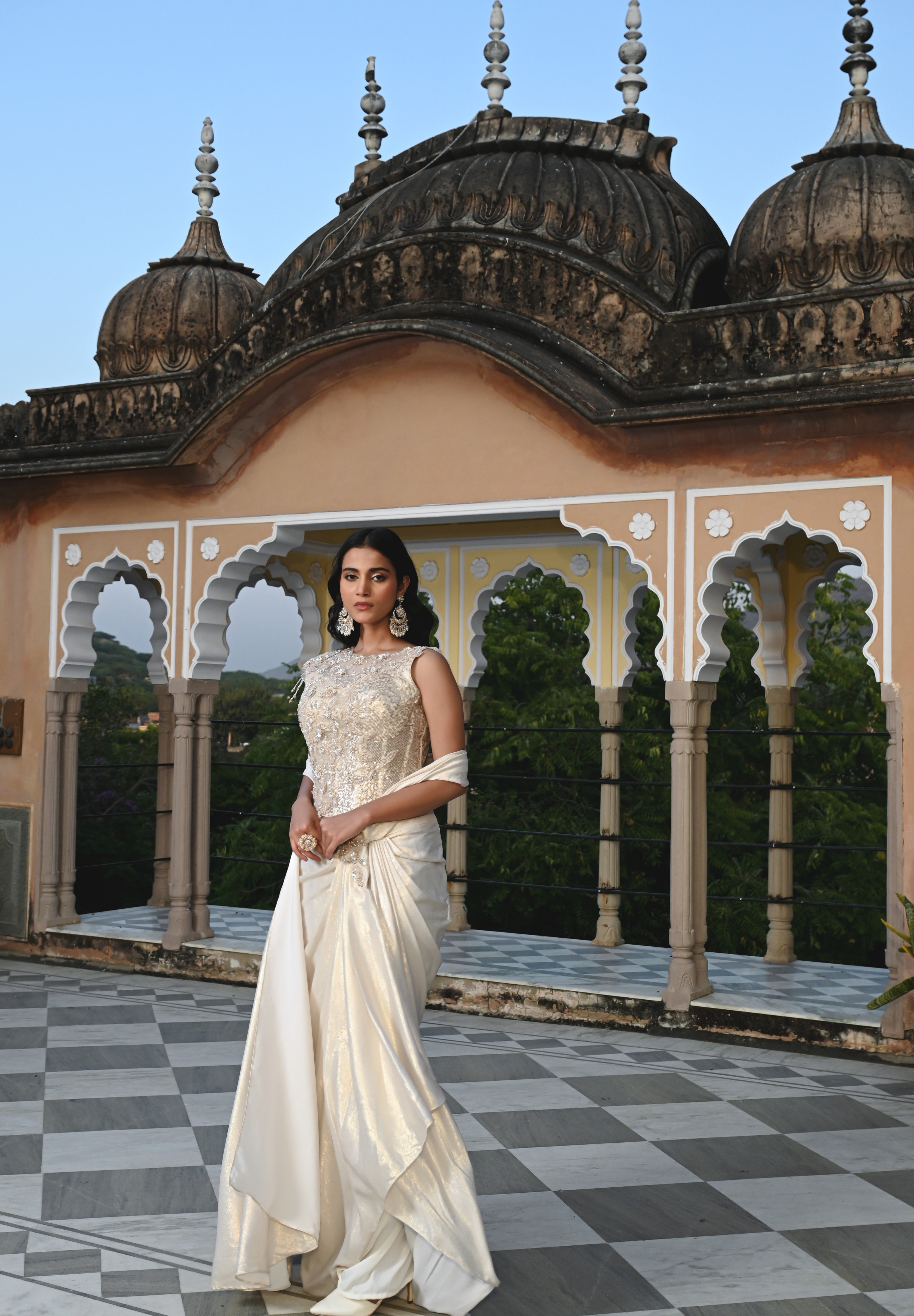 A woman poses on a rooftop in a shimmering ivory gown with a heavily embroidered bodice and a long, flowing train. The background features traditional Indian architectural elements like stone domes and scalloped arches.