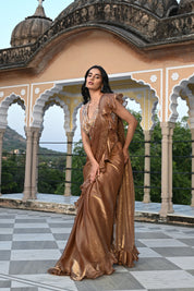 A woman poses on a rooftop terrace wearing a metallic bronze-toned draped saree featuring a voluminous ruffled border. The ensemble includes a sleeveless, sheer-style blouse heavily embellished with gold and silver floral embroidery. She stands before a backdrop of traditional white scalloped arches and lush green trees in the distance.