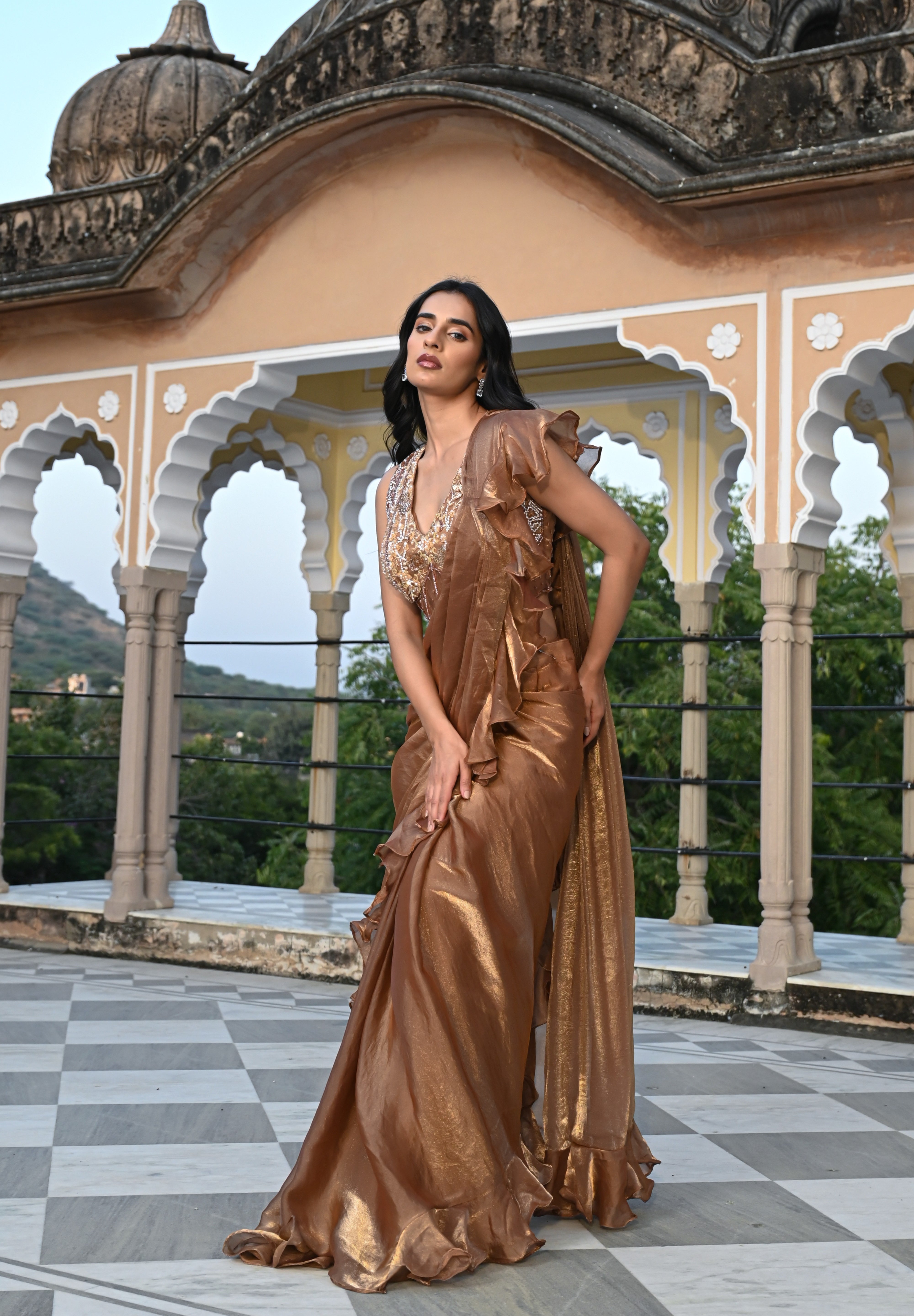 A woman poses on a rooftop terrace wearing a metallic bronze-toned draped saree featuring a voluminous ruffled border. The ensemble includes a sleeveless, sheer-style blouse heavily embellished with gold and silver floral embroidery. She stands before a backdrop of traditional white scalloped arches and lush green trees in the distance.